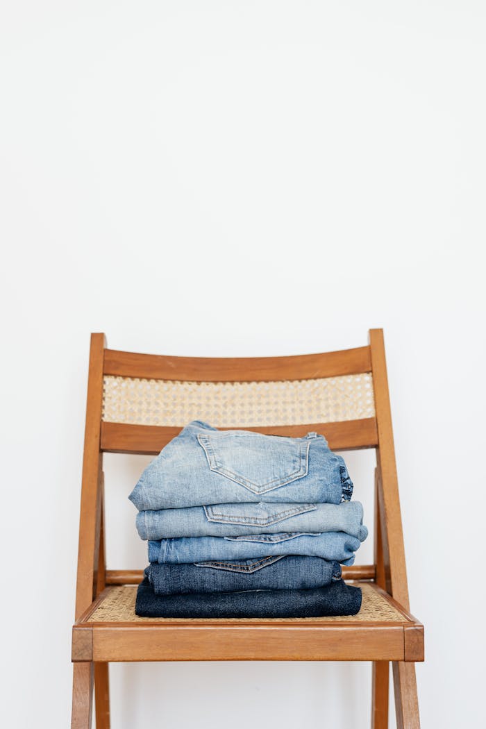 Stack of blue jeans of different shades on modern wooden chair against white wall